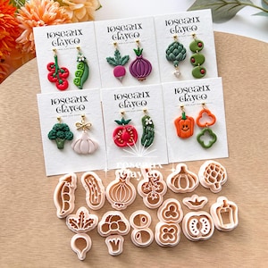 Puede incluir: Una colección de pendientes y cortadores de galletas de arcilla polimérica hechos a mano. Los pendientes presentan varios diseños de verduras, incluyendo tomates, guisantes, rábanos, cebollas, alcachofas, aceitunas, brócoli, ajo, pepinos y pimientos. Los cortadores de galletas tienen las mismas formas de verduras.