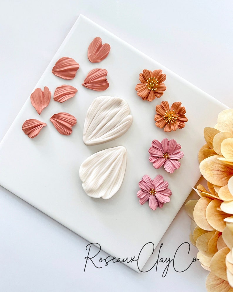 May include: A set of polymer clay flower petals and flowers in shades of pink, orange, and white. The petals are arranged on a white surface with the flowers below. The text "Roseaux Clay Co." is visible in the bottom right corner.