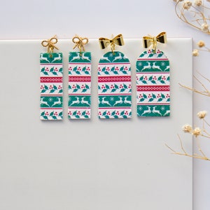 May include: Three pairs of green, red and white Christmas-themed earrings with gold bows. The earrings are rectangular and feature a repeating pattern of reindeer, holly and snowflakes.