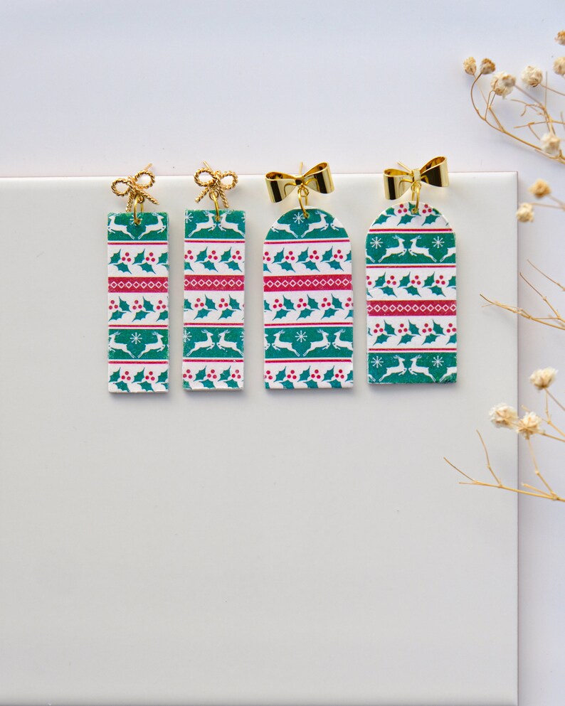 May include: Three pairs of green, red and white Christmas-themed earrings with gold bows. The earrings are rectangular and feature a repeating pattern of reindeer, holly and snowflakes.
