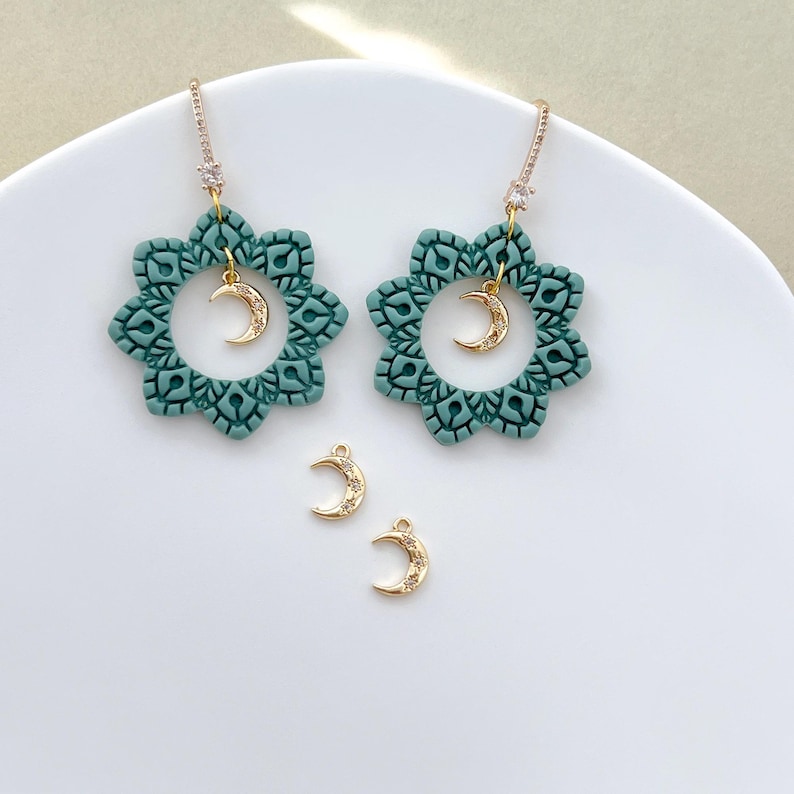 May include: Teal-colored floral earrings with gold-colored crescent moon charms. The earrings have a detailed, openwork design and are displayed on a white surface. Two additional crescent moon charms are also visible.