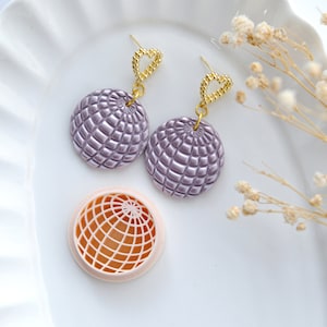 May include: A pair of purple and gold earrings with a heart-shaped design. The earrings are made of polymer clay and have a textured surface. The earrings are shown on a white plate with a small, pink, round mould.