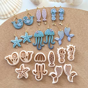 Puede incluir: Un conjunto de 16 pendientes y cortadores de galletas de arcilla polimérica con temas de vida marina. Los pendientes son en tonos azules y morados, mientras que los cortadores de galletas son de color marrón claro. Los pendientes incluyen caballitos de mar, medusas, estrellas de mar y focas. Los cortadores de galletas incluyen caballitos de mar, medusas, estrellas de mar, focas, sirenas y conchas.