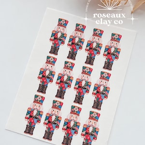 May include: A white rectangular sheet featuring twelve identical nutcracker illustrations arranged in three rows of four. Each nutcracker wears a red, blue, and green patchwork-style uniform with a matching hat. The text roseaux clay co is visible.