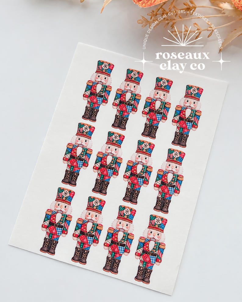 May include: A white rectangular sheet featuring twelve identical nutcracker illustrations arranged in three rows of four. Each nutcracker wears a red, blue, and green patchwork-style uniform with a matching hat. The text roseaux clay co is visible.
