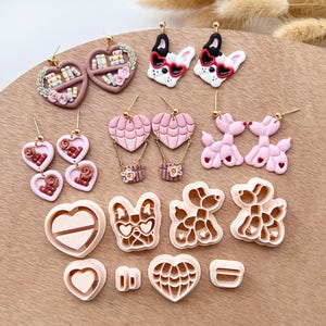 Op de afbeelding: Een set van 12 polymer clay oorbellen en uitstekers in roze en bruin. De oorbellen hebben verschillende ontwerpen, waaronder harten, boeken, luchtballonnen en honden. De uitstekers zijn in de vorm van harten, honden en luchtballonnen.
