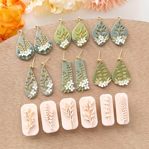Puede incluir: Un conjunto de 10 pendientes de arcilla polimérica de color verde y dorado con hojas y flores blancas y detalles dorados. Los pendientes se muestran sobre una superficie de madera con cuatro moldes de arcilla polimérica rosa para hacer los pendientes.