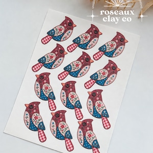 May include: A sheet of eleven bird-shaped cutouts featuring a red, blue, and white floral and gingham pattern. Each bird has a small black button eye. The sheet includes the text Roseaux Clay Co at the top right corner.