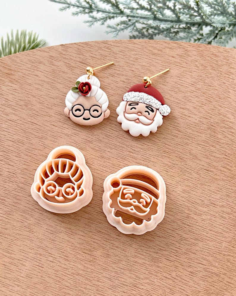 May include: A set of four Christmas-themed earrings and cookie cutters. The earrings feature Santa Claus and a woman with a red flower in her hair. The cookie cutters are also of Santa Claus and the woman.