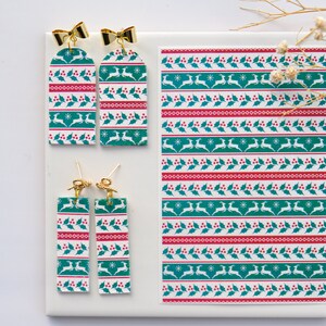 May include: A pair of dangle earrings with a green and white Christmas pattern featuring reindeer, holly, and snowflakes. The earrings are made of polymer clay and have gold-coloured hooks.