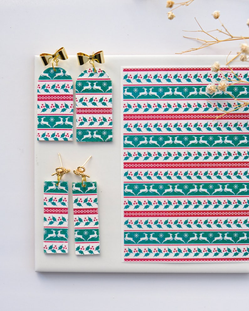 May include: A pair of dangle earrings with a green and white Christmas pattern featuring reindeer, holly, and snowflakes. The earrings are made of polymer clay and have gold-coloured hooks.
