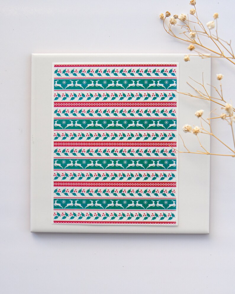 May include: A sheet of Christmas themed washi tape with a repeating pattern of green holly leaves, red berries, white snowflakes, and white reindeer.