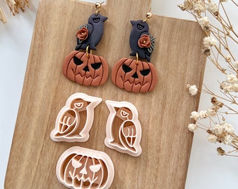 May include: A set of Halloween-themed earrings featuring black birds perched on top of orange pumpkins. The earrings are displayed on a wooden surface with two cookie cutters, one of a bird and one of a pumpkin.