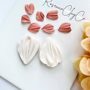 May include: A set of eight polymer clay flower petals in two colors: six pink petals and two white petals. The petals are arranged on a white surface with the text "Roseaux Clay Co." written in black cursive.