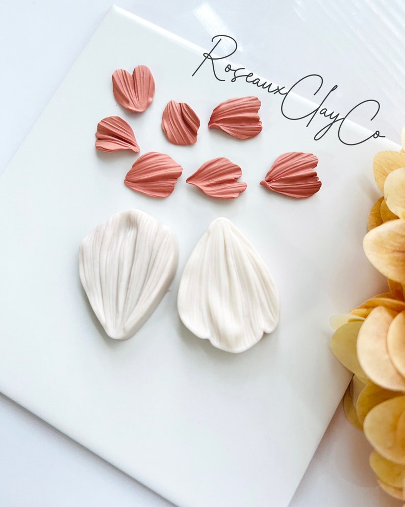 May include: A set of eight polymer clay flower petals in two colors: six pink petals and two white petals. The petals are arranged on a white surface with the text "Roseaux Clay Co." written in black cursive.