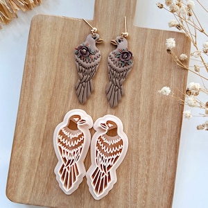 May include: Two pairs of brown bird earrings with floral accents and two white bird cookie cutters on a wooden surface.