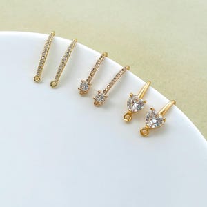 Puede incluir: Componentes de pendientes dorados con piedras de circonita cúbica transparentes y brillantes. Los componentes incluyen piezas en forma de barra con piedras incrustadas, gotas de una sola piedra y gotas en forma de corazón. Los pendientes se muestran sobre una superficie blanca.