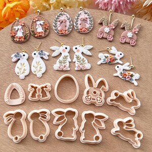 May include: A set of 14 polymer clay bunny and egg earrings and cookie cutters. The earrings are in pink, white, and gold tones. The cookie cutters are in a light brown color.