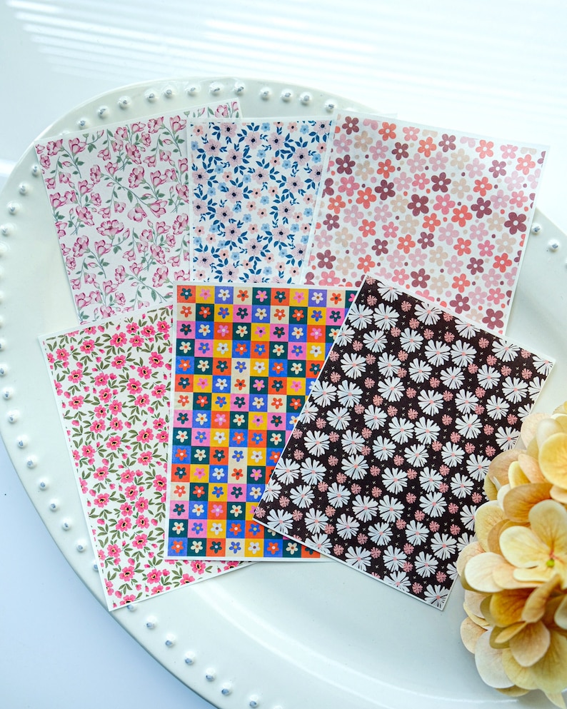 May include: Six different patterned sheets of paper with floral and geometric designs. The patterns include pink flowers on a white background, blue flowers on a white background, pink and red flowers on a white background, colourful squares with flowers on a yellow background, white flowers on a brown background, and pink flowers on a white background.