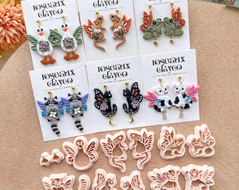 Könnte beinhalten: Handgefertigte Polymer Clay Ohrringe mit verschiedenen Tiermotiven, darunter Waschbären, Schmetterlinge und Kühe, auf weißen Karten präsentiert. Die Ohrringe zeigen lebendige Farben und aufwendige Details, zusammen mit passenden Ton-Ausstechern.
