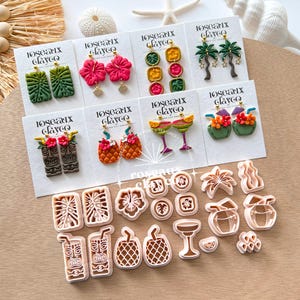 Op de afbeelding: Een verzameling oorbellen en kleisnijders met tropische thema's. De oorbellen hebben ontwerpen zoals hibiscusbloemen, palmbomen en tiki-maskers. De kleisnijders bevatten vormen van bloemen, ananassen en cocktailglazen. De oorbellen staan op witte kaarten met de tekst "loseaux clayco".