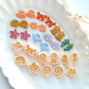 May include: A collection of 20 colourful polymer clay sea creature charms, including starfish, seashells, octopuses, crabs, and seahorses. The charms are arranged on a white plate.