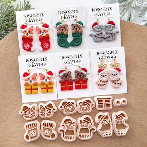 Puede incluir: Una colección de pendientes de arcilla polimérica hechos a mano con diseños festivos. Los pendientes incluyen corgis en calcetines, osos en calcetines, gatos grises, corgis en cajas de regalo, osos en cajas de regalo y llamas. También se muestran cortadores de galletas a juego.
