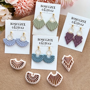 May include: An assortment of polymer clay earrings in various colors, including blue, green, and purple, displayed on white cards. The earrings feature gold-colored hoops and fan or geometric designs. The brand name "Roseaux Clayco" is printed on the cards.