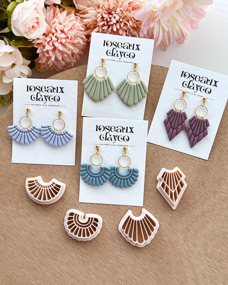 May include: An assortment of polymer clay earrings in various colors, including blue, green, and purple, displayed on white cards. The earrings feature gold-colored hoops and fan or geometric designs. The brand name "Roseaux Clayco" is printed on the cards.