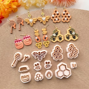 May include: A collection of 16 honey-themed polymer clay earrings and cookie cutters. The earrings feature bees, honeycombs, honey jars, and flowers. The cookie cutters are shaped like bees, honeycombs, and honey jars. The items are arranged on a wooden surface.