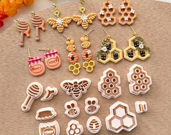 May include: A collection of 16 honey-themed polymer clay earrings and cookie cutters. The earrings feature bees, honeycombs, honey jars, and flowers. The cookie cutters are shaped like bees, honeycombs, and honey jars. The items are arranged on a wooden surface.