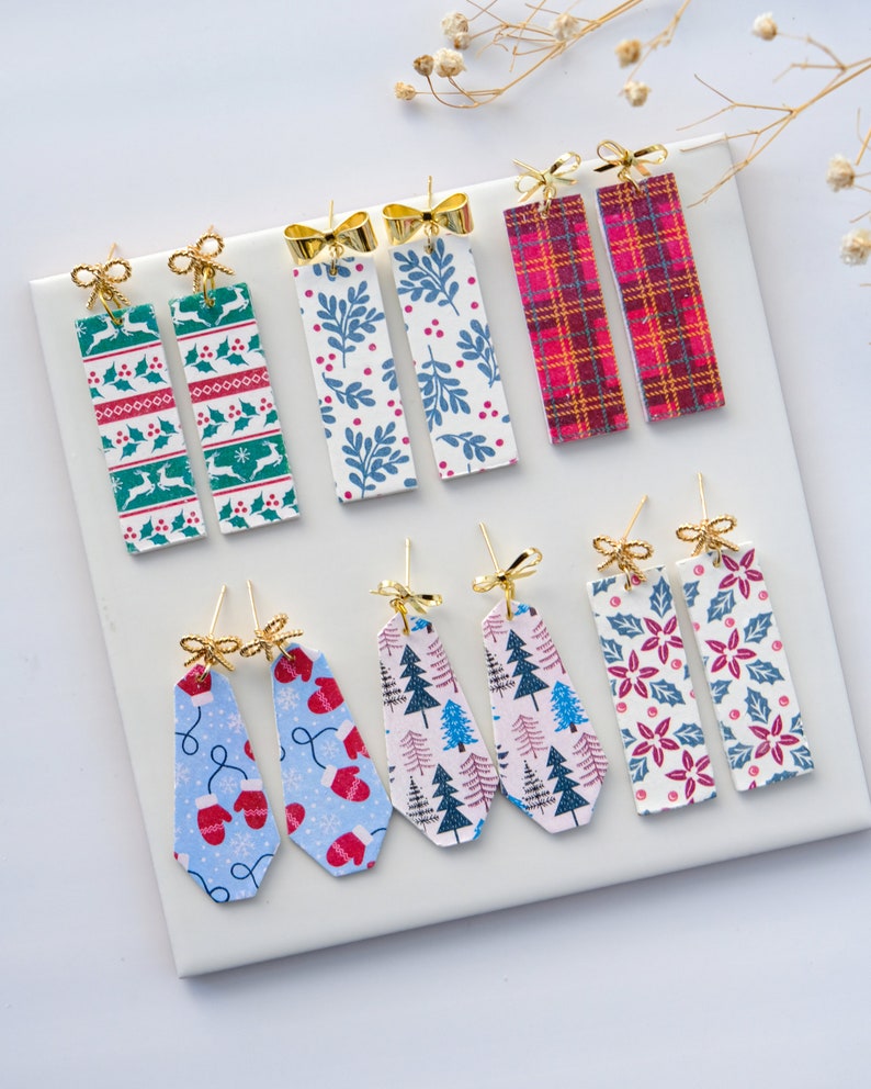 May include: A selection of Christmas-themed earrings featuring various patterns, including plaid, snowflakes, holly, and trees. The earrings are made with gold-coloured metal and have a bow detail.
