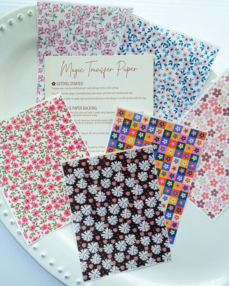 May include: A set of six sheets of floral patterned transfer paper for use with clay. The paper features a variety of floral designs, including pink flowers, white flowers, and colourful squares. The paper is labelled "Magic Transfer Paper" and includes instructions for use.