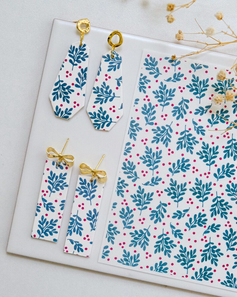 May include: A pair of dangle earrings and a pair of stud earrings with a white background and a blue and pink floral pattern. The earrings are made of polymer clay and have gold accents.