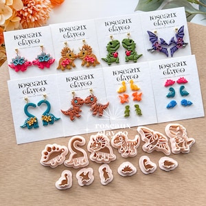 May include: A selection of polymer clay earrings and dinosaur-shaped cookie cutters. The earrings showcase diverse dinosaur designs, such as T-Rex, Triceratops, and Pterodactyls, with floral details. The cookie cutters are in various dinosaur shapes.