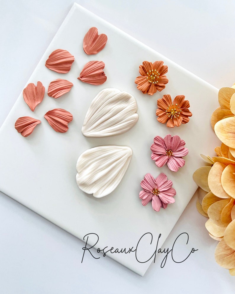 May include: A set of polymer clay flower petals and flowers in shades of pink, orange, and white. The petals are arranged on a white surface with the text "Roseaux Clay Co" in black script.