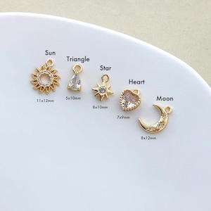 May include: A collection of gold-colored charms in various shapes: a sun (11x12mm), a triangle, a star (8x10mm), a heart (7x9mm), and a crescent moon (8x12mm). Each charm has a loop for attaching to jewelry. The charms are displayed on a white surface.