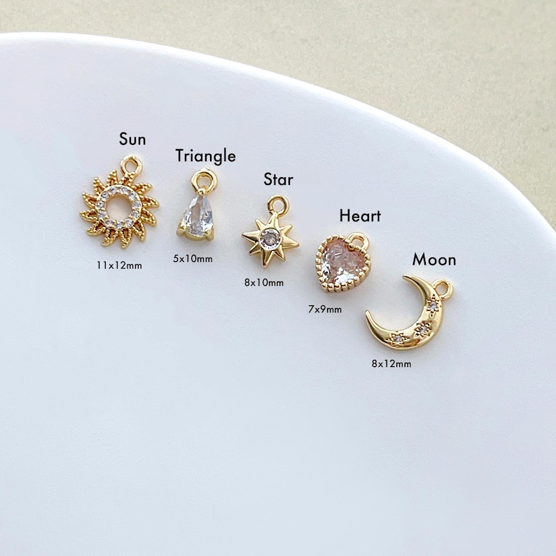 May include: A collection of gold-colored charms in various shapes: a sun (11x12mm), a triangle, a star (8x10mm), a heart (7x9mm), and a crescent moon (8x12mm). Each charm has a loop for attaching to jewelry. The charms are displayed on a white surface.