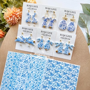 Può includere: Una collezione di orecchini con motivi floreali blu e bianchi in vari design, tra cui forme di coniglietti e gocce, esposti su cartoncini bianchi. Gli orecchini hanno dettagli color oro e sono disposti su uno sfondo di legno. Il marchio "Roseaux Clayco" è visibile.