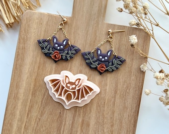 May include: A pair of gold dangle earrings featuring a purple bat charm and a small orange flower. The bat is surrounded by green leaves. The earrings are displayed on a wooden surface with a pink bat cookie cutter.