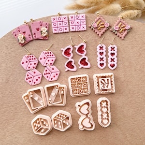 Könnte beinhalten: Ein Set mit 12 rosafarbenen und goldenen Ohrringen mit verschiedenen Designs zum Thema Spiele, darunter Würfel, Spielkarten, Tic-Tac-Toe und Herzen. Die Ohrringe werden auf einer Holzoberfläche präsentiert.