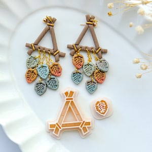 May include: A pair of dangle earrings with a triangular frame made of brown clay and gold chains with small clay leaves in shades of green, orange, and brown hanging from the bottom.