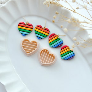 May include: Four rainbow heart-shaped earrings with gold posts, two of the earrings are on a white plate and two are in their heart-shaped molds.