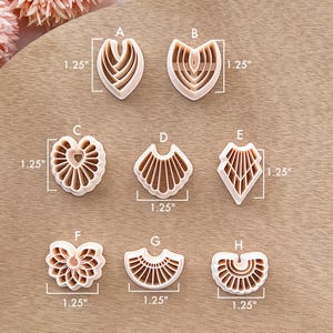 May include: A collection of eight beige polymer clay cutters in various shapes, each measuring 1.25 inches. Designs include heart, fan, and geometric patterns, ideal for crafting. The cutters are arranged on a light brown wooden surface.