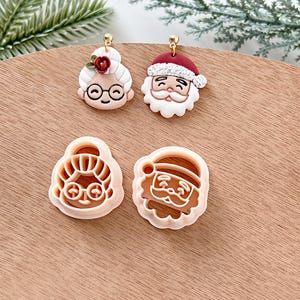 May include: Two pairs of Christmas-themed cookie cutters, one of Santa Claus and one of a woman with glasses and a flower in her hair. The cutters are made of white plastic and are on a wooden surface.