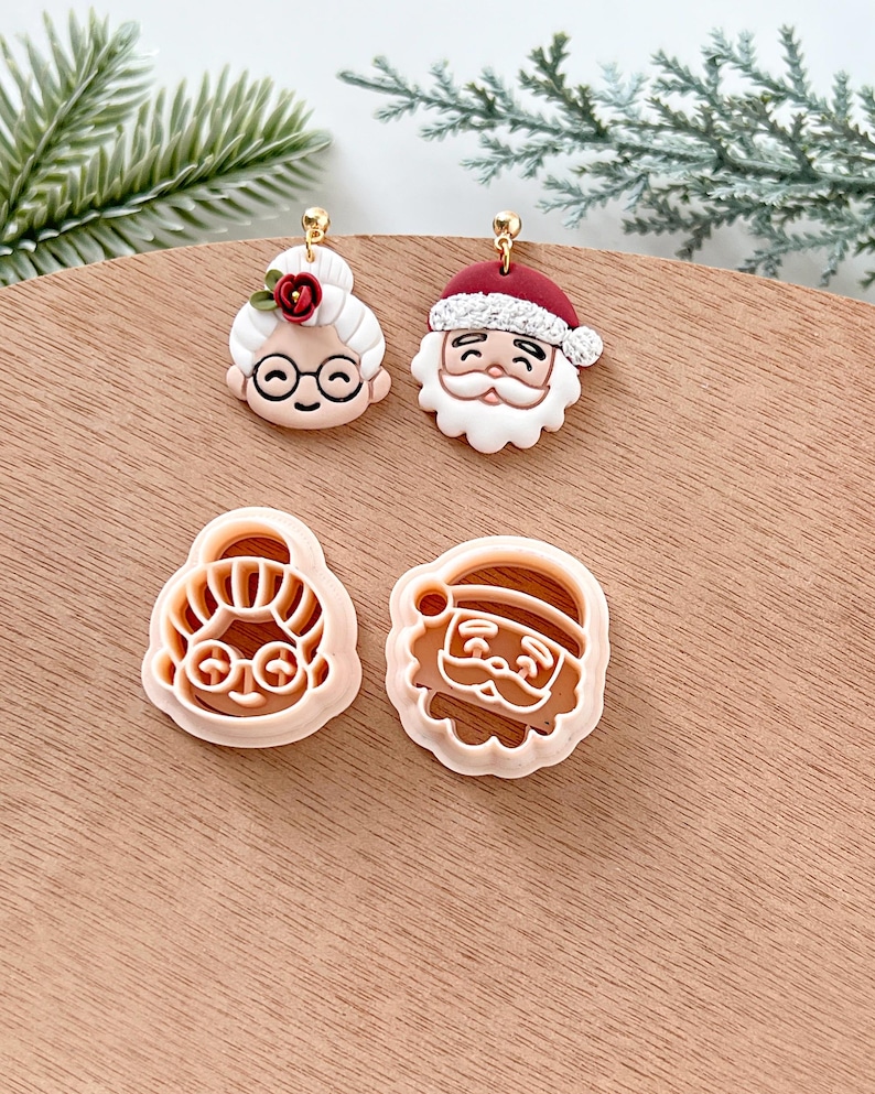 May include: Two pairs of Christmas-themed cookie cutters, one of Santa Claus and one of a woman with glasses and a flower in her hair. The cutters are made of white plastic and are on a wooden surface.