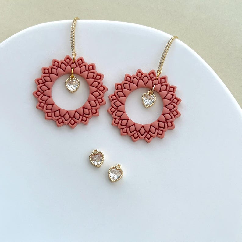 May include: Two pairs of earrings. The first pair features large, dusty rose-colored, circular pendants with a floral design. Each pendant has a small, heart-shaped, clear crystal charm. The second pair of earrings are small, heart-shaped, clear crystal charms with gold-colored settings.