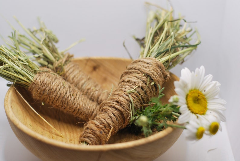 May include: Two bundles of dried grass wrapped in twine, sitting in a wooden bowl with a white daisy flower.