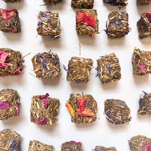Op de afbeelding: Een verzameling van 25 kleine, vierkante gedroogde bloemen- en kruidenblokjes. De blokjes zijn een mix van groen, bruin, roze, paars en oranje.