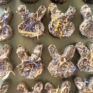 Puede incluir: Una colección de golosinas en forma de conejo hechas a mano con hierbas y flores secas. Las golosinas son de color marrón claro y tienen varias flores moradas y amarillas incrustadas.